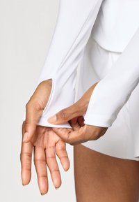 Balta ilgomis rankovėmis palaidinė su pirštų anga rankogalyje, minkšta medžiaga, prigludusi forma, glotnus paviršius ir šiek tiek elastinga.