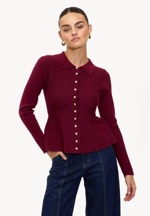 Haut en maille bordeaux avec col, présentant des boutons dorés sur le devant, manches longues et un ourlet à basque. Associé à un denim taille haute.