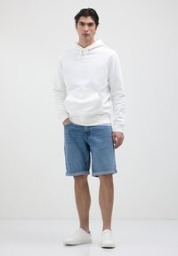 Haut à capuche en coton blanc avec une poche avant, associé à un short en denim bleu clair avec des manches retroussées et des baskets blanches. Tenue simple et décontractée.