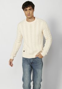 Suéter de punto cableado crema con escote redondo y puños acanalados, combinado con jeans de mezclilla azul. Detalle de marca sutil en el dobladillo.