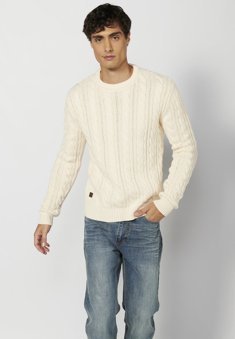 Suéter de punto cableado crema con escote redondo y puños acanalados, combinado con jeans de mezclilla azul. Detalle de marca sutil en el dobladillo.
