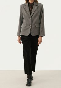 Femme marchant portant un blazer gris à deux boutons, un haut en dentelle noire, un pantalon en velours côtelé noir, des collants noirs et des mocassins noirs.