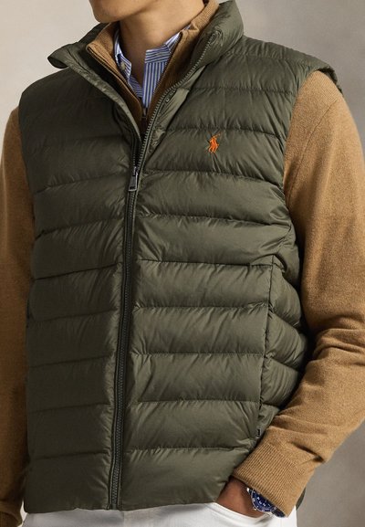 Olivengrønn puffvest med horisontal quilting, glidelåslukking og oransje logodetalj, lagvis over en tanfarget genser med ribbestrikkede mansjetter.