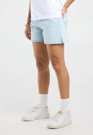Persoon draagt lichtblauwe denim short, witte sokken, witte New Balance sneakers en een wit bovenstuk, staand tegen een effen achtergrond.
