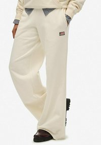 Persoon in een crème-kleurige wijde broek met een klein rechthoekig vlaggenpatch, gecombineerd met donkerbruine schoenen en handen in de zakken.