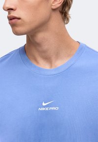 Νεαρό άτομο με ανοιχτό γαλάζιο μπλουζάκι Nike Pro, που δείχνει το ντεκολτέ και το μερικό πρόσωπο στραμμένο αριστερά σε απλό φόντο.