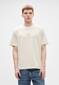Μπεζ βαμβακερό T-shirt με στρογγυλή λαιμόκοψη και κοντά μανίκια, που διαθέτει κεντημένο λογότυπο CK στο μπροστινό μέρος. Φοριέται με μπλε τζιν παντελόνι.