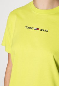 Kirkas keltainen puuvillainen t-paita, jossa on lyhyet hihat. Paidassa on mustalla brodeerattu "TOMMY" ja "JEANS" vaakasuoran punaisen, valkoisen ja sinisen raidankin yläpuolella.