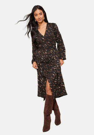 Schwarzes Kleid mit abstraktem braun-beigem Muster, langen Ärmeln, V-Ausschnitt, Knopfleiste vorne und Seitenschlitz; kombiniert mit braunen kniehohen Stiefeln.