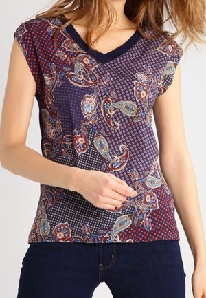 Top sans manches à col en V avec des motifs mixtes paisley et floraux en bleu, rouge et beige, porté avec un jean bleu foncé.