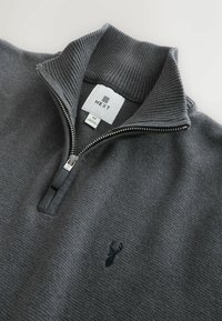 Grijze zip-up trui van textuurstof, met een ribbelkraag en een klein geborduurd logo op de borst. Maatlabel: M.