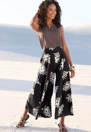 Top marrone senza maniche abbinato a pantaloni larghi neri con motivi floreali bianchi. Il modello indossa sandali neri e bracciali impilati.