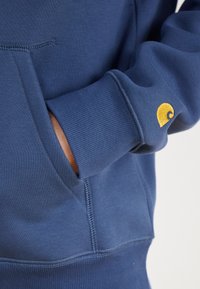 Navy blue sweatshirt featuring a textured fabric, ribbed cuffs, and a small yellow embroidered logo on the left sleeve.
