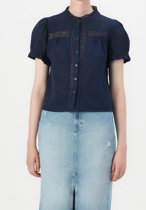 Femme portant un chemisier bleu marine à manches courtes avec des détails en dentelle et des boutons, associé à une jupe en jean bleu clair avec une fente à l'avant.