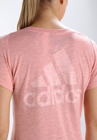 Světle růžové sportovní tričko vyrobené z jemné, texturované látky. Na zádech se nachází velké, bledé logo Adidas. Krátké rukávy a kulatý výstřih.