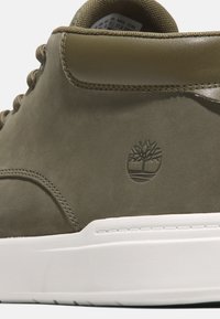 Sapatilhas de couro verde-azeitona com sola de borracha branca, apresentando um acabamento texturizado e um design de logótipo em relevo na lateral.