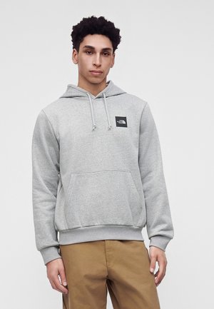 Jeune homme portant un sweat à capuche gris clair avec une poche avant et un logo, associé à un pantalon marron clair, debout devant un fond uni.