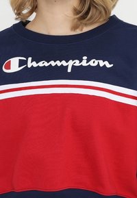 Tmavě modrá a červená bavlněná mikina s bílými pruhy, s bílým logem "Champion" napříč hrudníkem. Krátké rukávy, design s kulatým výstřihem.