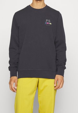 Mann trägt einen dunkelgrauen Sweatshirt mit langen Ärmeln und kleinem buntem Text auf der Brust und leuchtend gelbe Hose, steht vor einem einfarbigen Hintergrund.