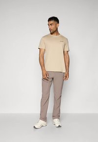 Muškarac u bež košulji s kratkim rukavima, svijetlosmešnim hlačama i bijelim sportskim cipela, stoji i gleda lijevo uz jednostavnu pozadinu.