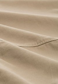 Beige Stoff mit einer glatten Textur, der gefaltete Lagen und einen subtilen Glanz aufweist. Sichtbare Nähte entlang der Kanten betonen die Details.