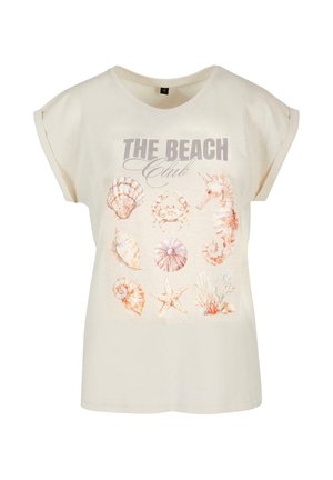 T-shirt en coton beige avec des manches roulées, arborant un motif sur le thème de la plage avec des coquillages et un hippocampe, ainsi que le texte "LE CLUB DE LA PLAGE."