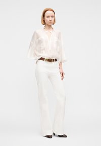 Mujer de pie que lleva una blusa blanca translúcida con mangas anchas, pantalones blancos de talle alto, cinturón marrón y zapatos oscuros de punta afilada.
