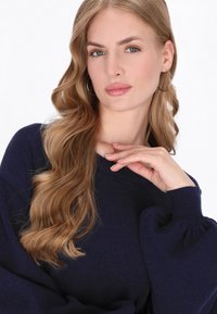 Granatowy sweter robiony na drutach z bufiastymi rękawami i okrągłym dekoltem. Widoczne długie, falowane włosy modelki oraz minimalne srebrne kolczyki w kształcie obręczy.