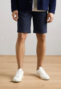 Marineblå shorts laget av bomull, med et enkelt design og rett snitt. Kombinert med hvite joggesko. Benene synlige, stående på tregulv.