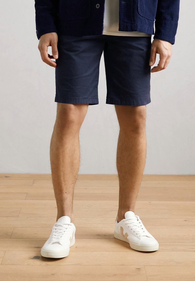 Marineblau Shorts aus Baumwolle, mit einem schlichten Design und geradem Schnitt. Kombiniert mit weißen Sneakers. Beine sichtbar, stehend auf einem Holzboden.
