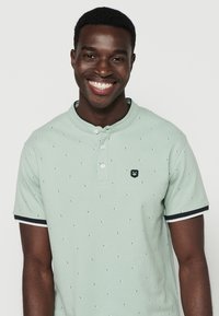Polo shirt verde chiaro con piccoli motivi, tre bottoni al colletto e accenti a strisce blu navy sulle maniche.