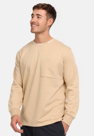 Camisa beige de manga larga con cuello redondo. Presenta un bolsillo en el pecho y un detalle de logo en el cuello. Textura suave con un ajuste relajado.
