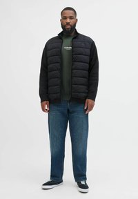 Schwarze wattierte Weste mit Reißverschluss vorne, kombiniert mit einem grünen Sweatshirt und blauen Jeans. Die Weste hat einen hohen Kragen und einen strukturierten Stoff.