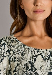 Unteres Gesicht und Nacken einer Frau, die ein Oberteil mit Schlangenhautmuster trägt, dazu neutrale Lippenfarbe und welliges braunes Haar.