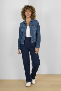 Chaqueta de mezclilla en azul medio con cierre de botones, bolsillos en el pecho y mangas largas. Combinada con jeans oscuros de cintura alta y zapatillas blancas.
