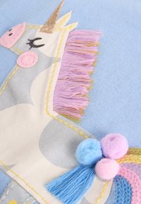 Décoration en peluche licorne avec un design gris et blanc, accents roses, corne dorée et franges pastel texturées. Fond bleu mettant en valeur les détails.