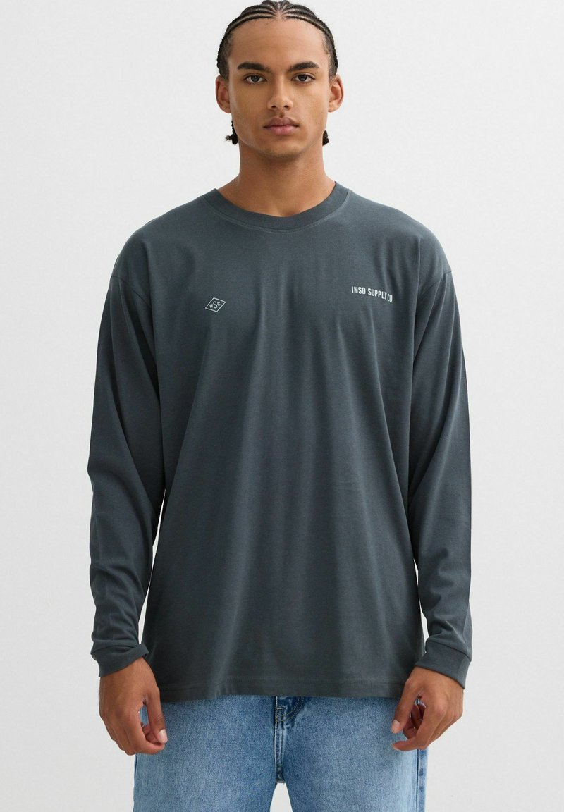 Camisa de manga larga de color gris oscuro, fabricada con un tejido suave, con un cuello redondeado y un pequeño logo blanco en el pecho. Combinada con vaqueros azules.