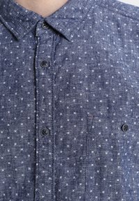 Een blauwe button-up shirt van textuurstof met een klein wit bloemenpatroon, een ronde kraag en twee voorzakken met donkere knopen.
