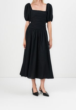 Frau trägt ein schwarzes Midikleid mit gepufften kurzen Ärmeln und gerafftem Oberteil, kombiniert mit schwarzen Slingback-Absätzen, stehend vor weißem Hintergrund.