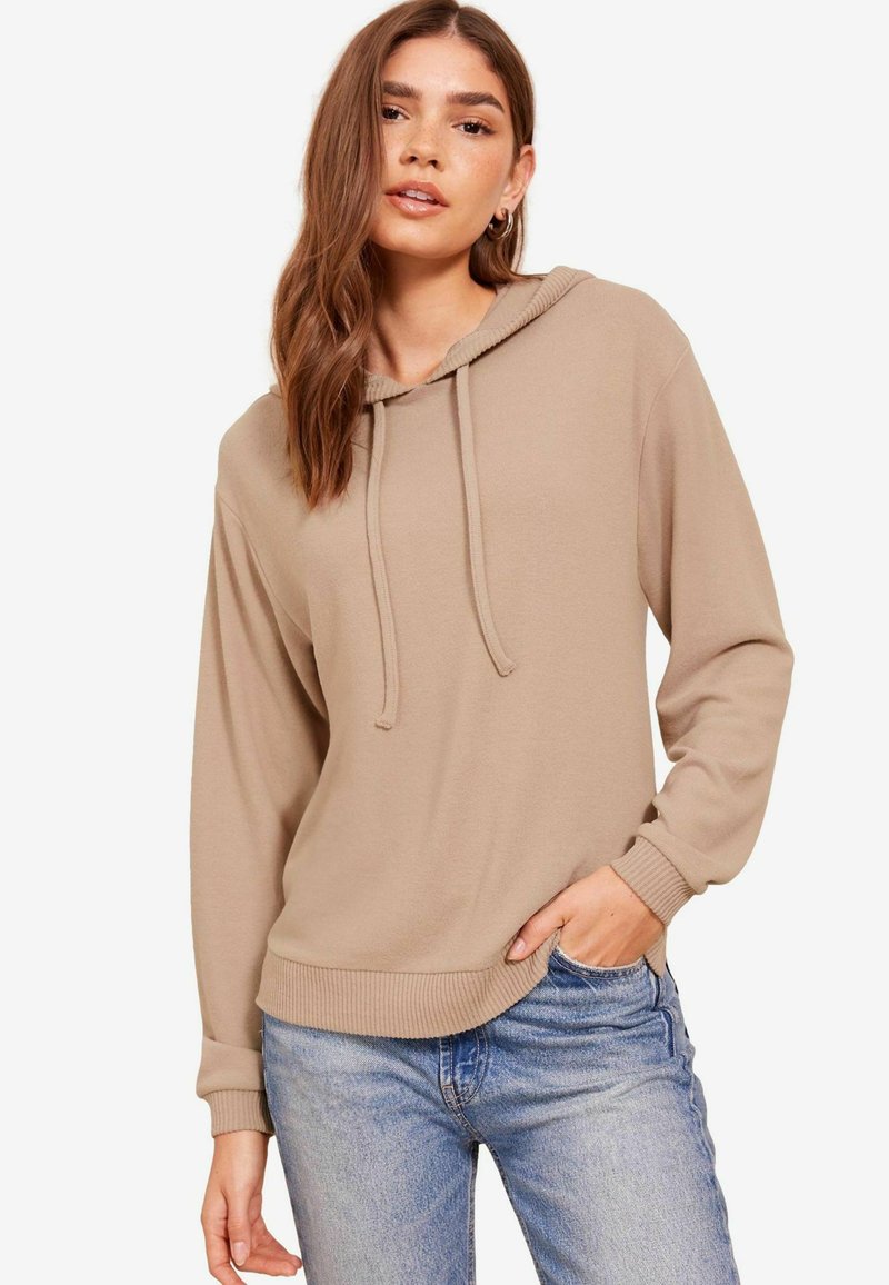 Felpa beige con orlo e polsini a coste, dotata di cappuccio con cordino. Il tessuto appare morbido con una vestibilità rilassata. Indossata con jeans blu.