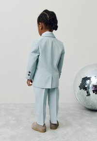 Otrok s spetimi kitkami, oblečen v svetlo modro obleko in rjave čevlje, stoji obrnjeno stran ob veliki srebrni disko krogli na sivem tleh.