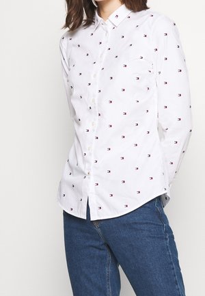 Femme portant une chemise blanche ajustée avec de petits motifs de logos rouges et noirs associée à un jean bleu, debout devant un fond uni.