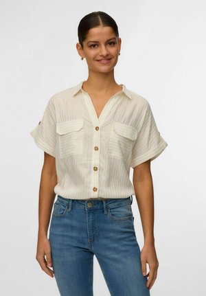 Femme aux cheveux foncés en chignon bas portant une chemise blanche à manches courtes à fines rayures et un jean bleu, souriant à la caméra.