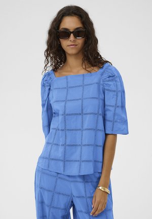 Kobieta o kręconych włosach, nosząca czarne okulary przeciwsłoneczne, niebieską kratowaną bluzkę i dopasowane spodnie oraz złotą bransoletkę na nadgarstku.