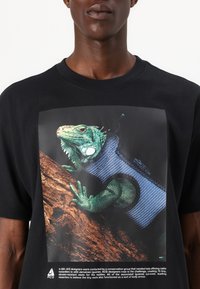 Sort T-shirt med et livligt grafisk tryk af en grøn firben iført en blå vest, sat mod en baggrund af naturligt træ.