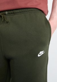 Nike olīvzaļas sporta bikses, kas izgatavotas no mīkstas auduma, ir aprīkotas ar elastīgu jostasvietu, auklu un baltu logo kreisajā apakšējā kājas daļā.