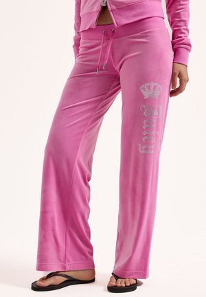 Rosafarbene Velourhosen mit Kordelzug in der Taille und Rhinestone-Schriftzug "Juicy" sowie Kronenlogo am linken Bein, getragen mit schwarzen Flip-Flops und passender Jacke.