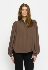 Brun, löst sittande blus med krage, knäppning fram och rynkade ärmar. Mjukt tyg med en slät yta. Bärs med svarta byxor.