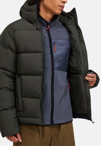 Polstret mørkegrøn jakke med hætte, med en lynlåset brystlomme i blå fleece og orange lynlås-dimser. Indre fleecelag synligt.