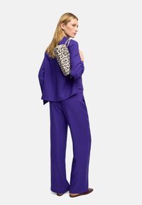 Fioletowy, fakturowany zestaw składający się z luźnych spodni i dopasowanej bluzki. Na ramieniu zawieszona jest w kolorze beżowym torba w kropki.